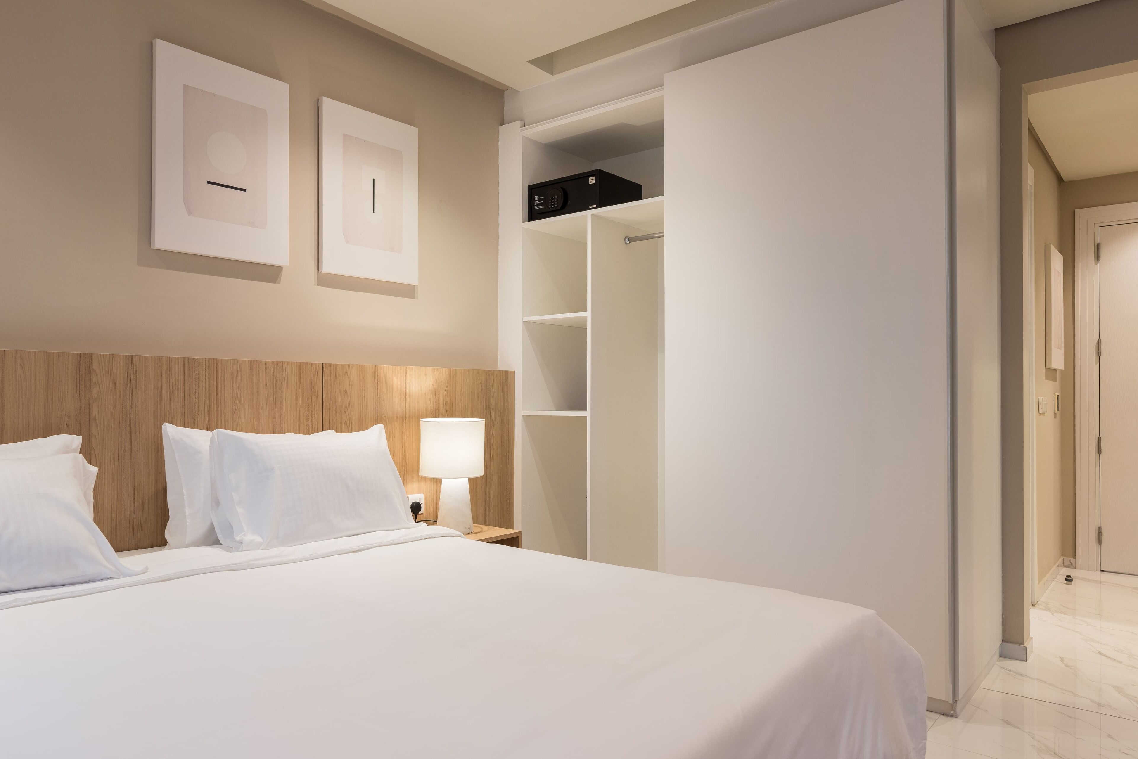 Habitación de hotel con una cama grande, cabecera de madera, armario blanco y mesita de noche con una lámpara.