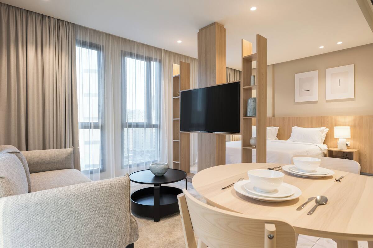 A hotel room with a sofa, a dining table set with bowls and plates, a flat-screen TV, and a bed with a nightstand.