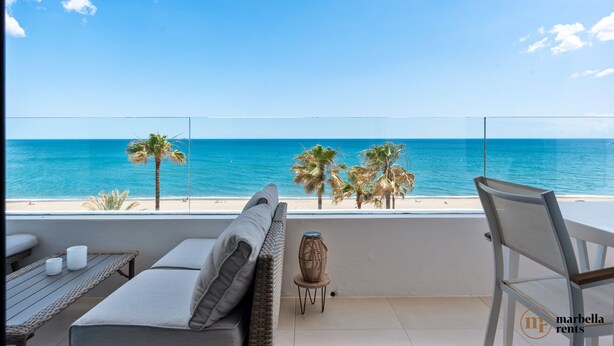 Un balcón con vista al mar, una silla de mimbre, una mesita con un florero y un sofá con cojines.