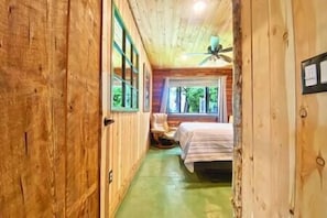Un étroit couloir en bois mène à une chambre qui contient un lit, une chaise et un ventilateur de plafond.