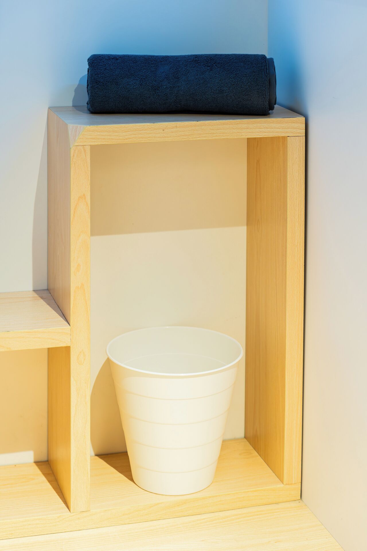 Ein Holzregal mit einem weißen Kunststoffkübel und einem dunkelblauen Handtuch darauf.