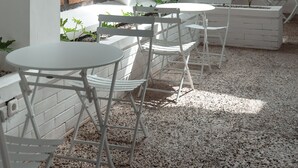 A white outdoor seating area with tables and chairs, a wooden shuttered window, and a white brick planter with plants. - Bobopod Kota Lama Semarang (Semarang)
