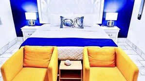 A hotel room with a bed, two bedside tables with lamps, a blue accent wall, and two yellow armchairs. - Sapphire by the Sea Unit 8 - King Pool View Suite, kitchen, pool view, Sleeps 2 (Fort Lauderdale)