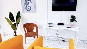 A modern living room with a white wall, a mounted TV, a wicker chair, a white desk, and a potted plant. - Sapphire by the Sea Unit 8 - King Pool View Suite, kitchen, pool view, Sleeps 2 (Fort Lauderdale)