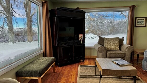A cozy living room with a wooden floor, a dark entertainment unit, a sofa with a patterned cushion, and a coffee table with a book. - Edge Water Lodge (EV), Waters Edge Lodge (Fremont County)