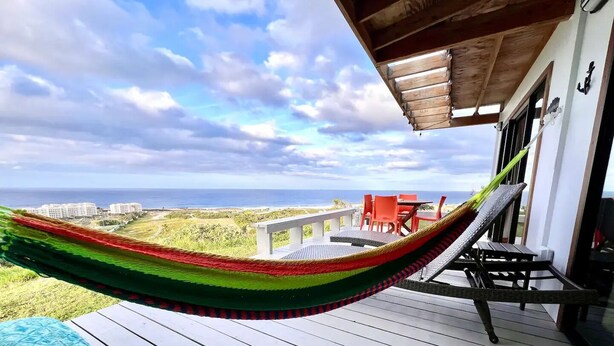 A hammock on a wooden deck with a view of the ocean and buildings in the background.
