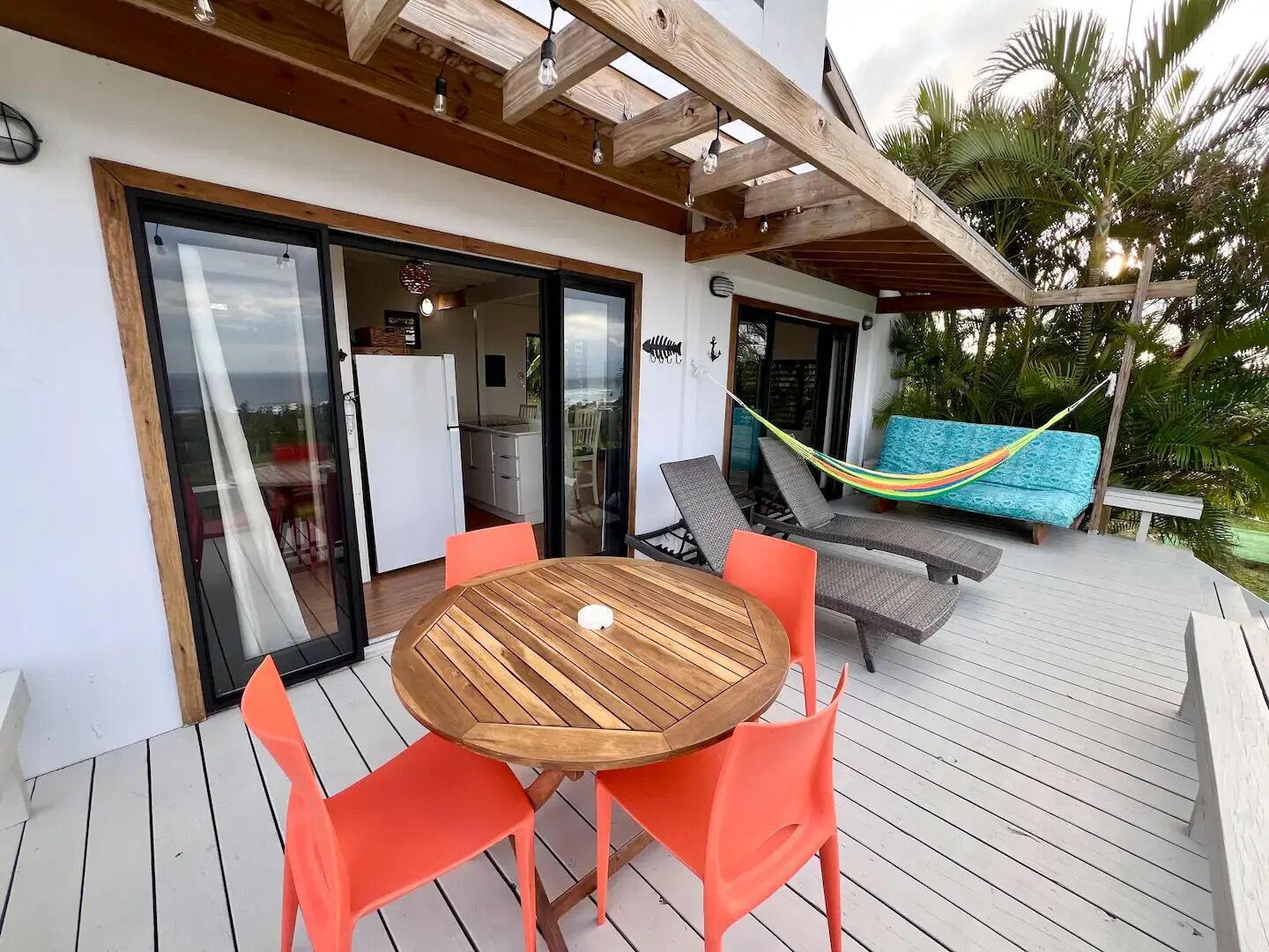 A wooden deck with a round table and orange chairs, a hammock, and a view of a kitchen and sliding glass doors.