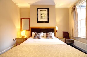 A hotel room with a large bed, a nightstand with a lamp, a chair, and a window with curtains. - Chester Apartments (Apt B) (Chester)
