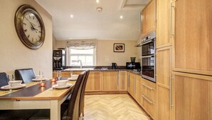 A kitchen with wooden cabinets, a dining table set for four, a clock on the wall, and a window with a view. - Chester Apartments (Apt B) (Chester)