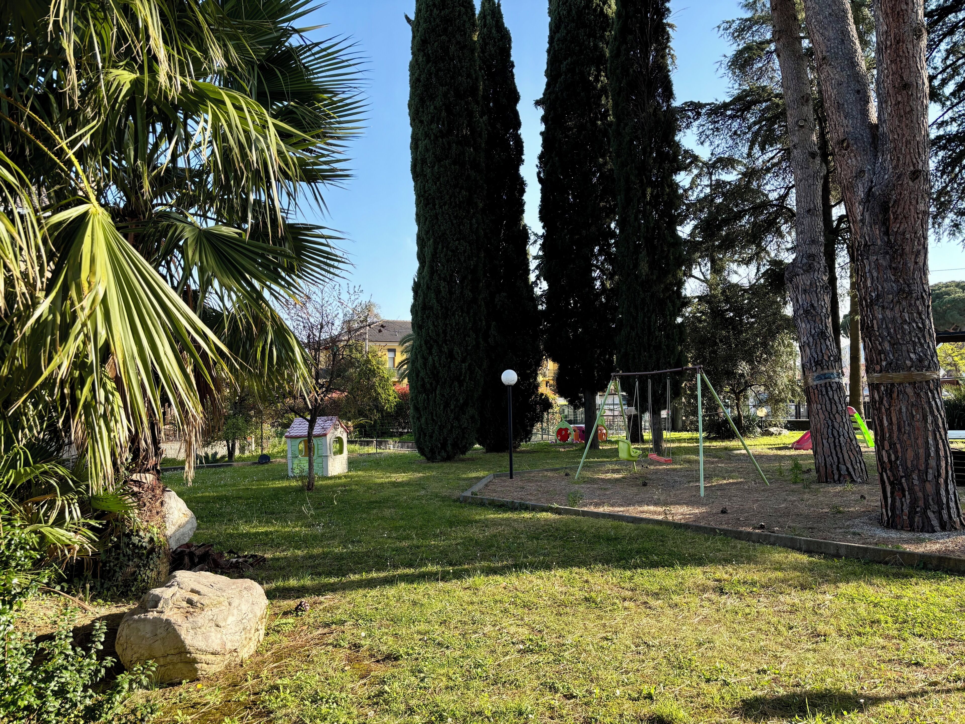 Egy park, amelyben hinta található, magas fák állnak, és egy kis játszótár is van.