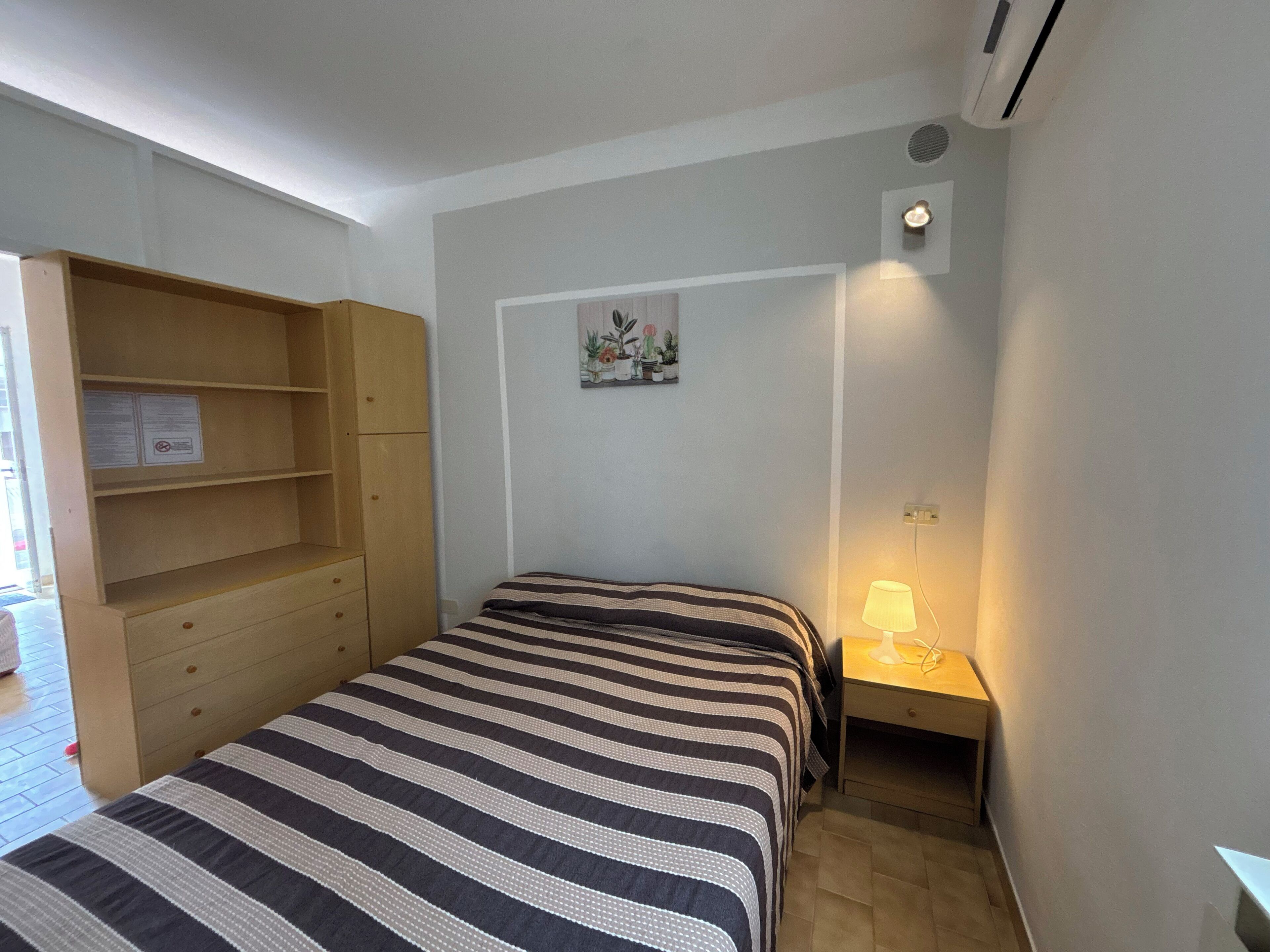 Un dormitorio con cama, un armario de madera, una mesita de noche con lámpara y un cuadro colgado en la pared.