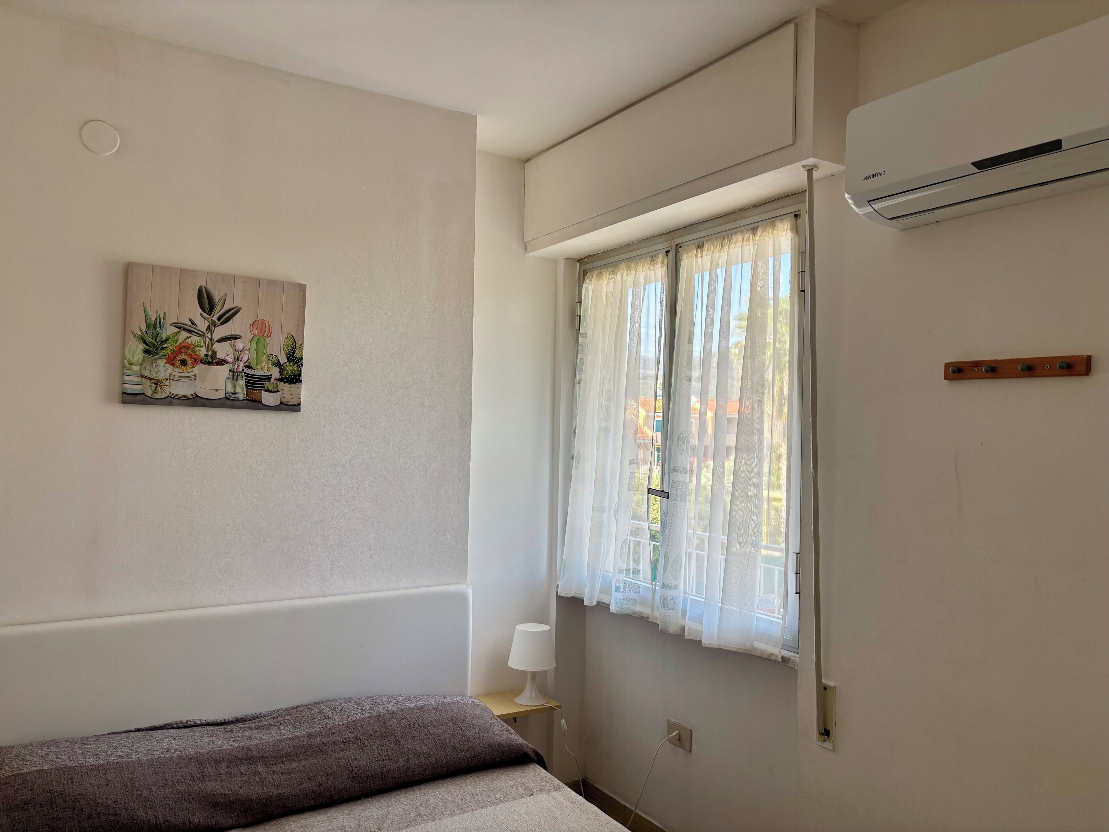 A bedroom with a bed, a window with curtains, an air conditioner, a wall-mounted lamp, and a painting.