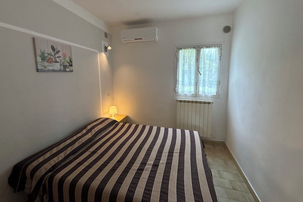 A room with a bed, a nightstand with a lamp, a radiator, a window with curtains, and a wall-mounted air conditioner.