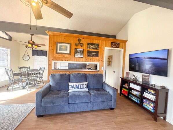 A living room with a gray sofa, wooden walls, a dining area, and a TV on the wall.