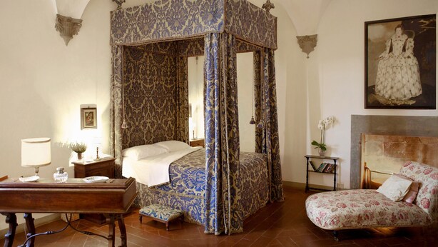 A four-poster canopy bed with a floral pattern, a wooden nightstand with a lamp, a chair with a floral cushion, and a painting on the wall. - Castello Lorenzo Heart of Tuscany Renaissance Villa With Pool (San Casciano in Val di Pesa)