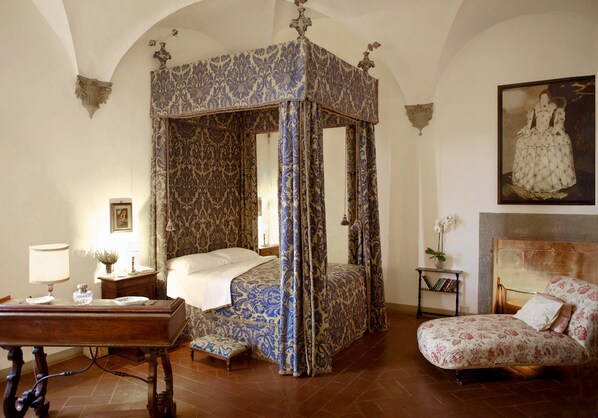 A four-poster canopy bed with a floral pattern, a wooden nightstand with a lamp, a chair with a floral cushion, and a painting on the wall.