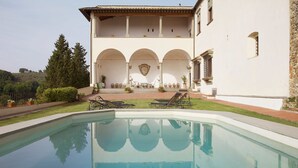 A pool area with lounge chairs and a dining table, surrounded by a building with arches and a view of the hills. - Castello Lorenzo Heart of Tuscany Renaissance Villa With Pool (San Casciano in Val di Pesa)