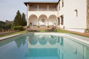 A pool area with lounge chairs and a dining table, surrounded by a building with arches and a view of the hills.