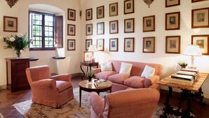 A cozy living room with a sofa, armchairs, a coffee table, and a bookshelf. There are framed pictures on the wall, a window with shutters, and a vase with flowers on a cabinet. - Castello Lorenzo Heart of Tuscany Renaissance Villa With Pool (San Casciano in Val di Pesa)