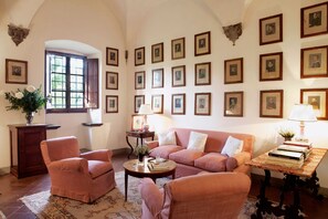 A cozy living room with a sofa, armchairs, a coffee table, and a bookshelf. There are framed pictures on the wall, a window with shutters, and a vase with flowers on a cabinet.