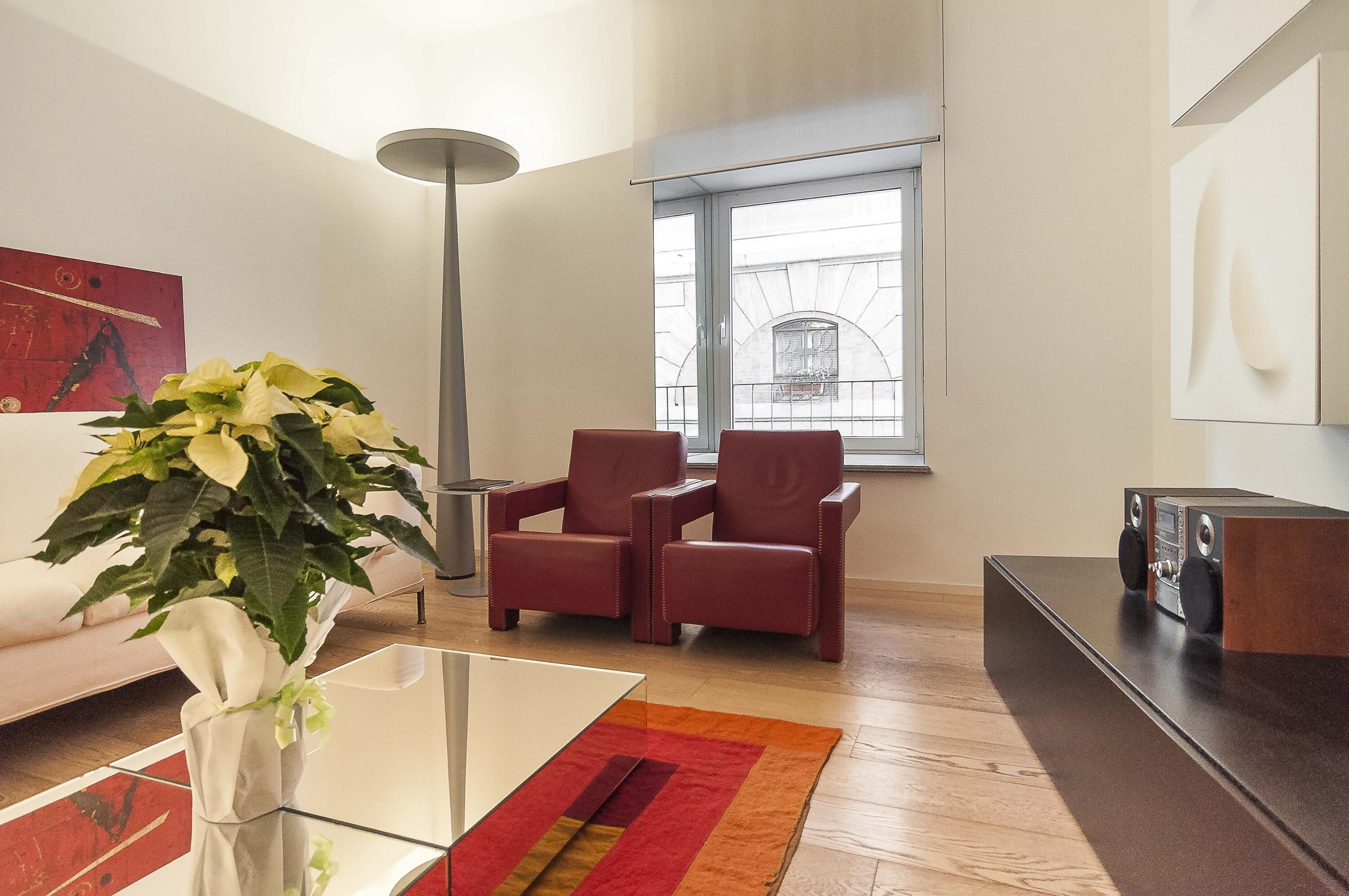 Una sala de estar moderna con un sofá, dos sillones rojos, una mesa de centro de vidrio, una lámpara de pie, un ventanal grande y un equipo de sonido.
