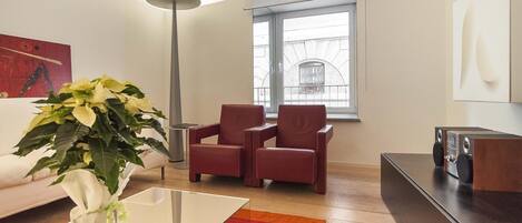 Una sala de estar moderna con un sofá, dos sillones rojos, una mesa de centro de vidrio, una lámpara de pie, un ventanal grande y un equipo de sonido.