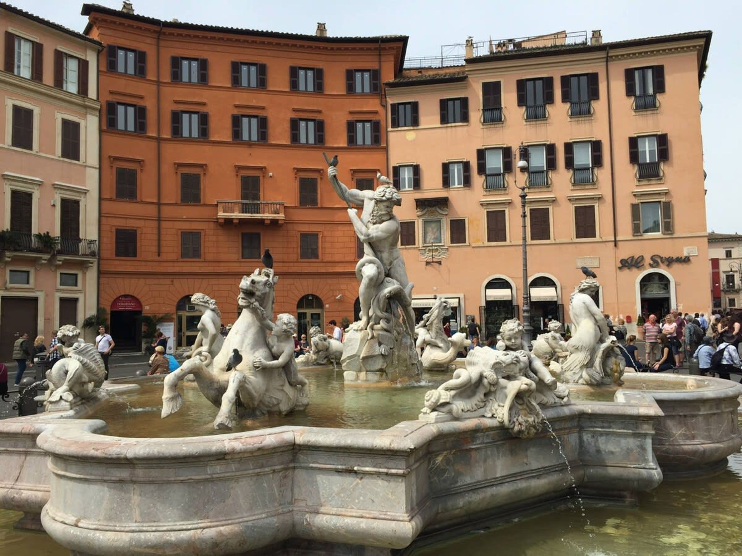 A historic fountain with classical sculptures in a public square, surrounded by multi-story buildings.