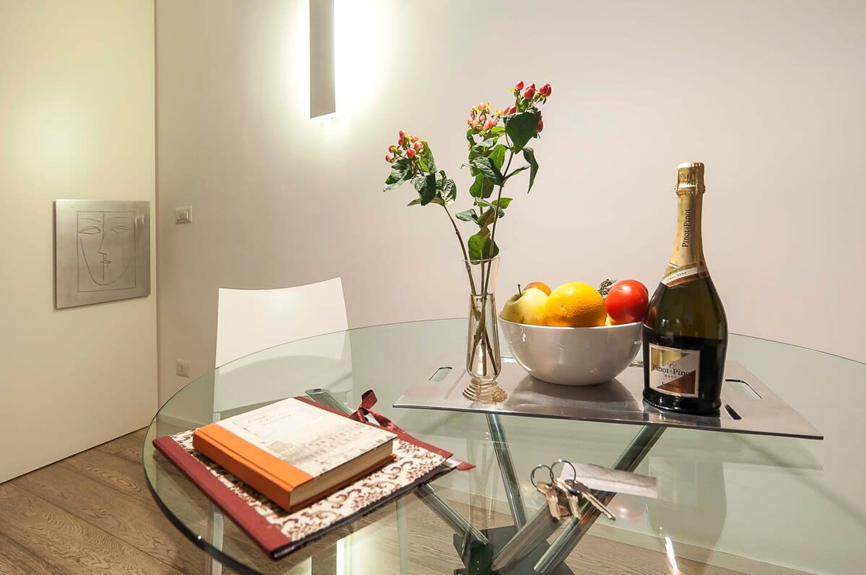 A glass table with a bowl of fruit, a bottle of wine, and a vase with flowers.