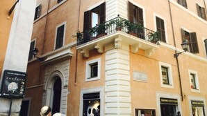 A multi-story building with a beige facade, white trim, and a central white column. There are storefronts on the ground floor with signs, and a few people are visible on the street. - In Rome at Spanish Steps Classy Apartment With Modern Design in an Historic Palazzo (Rome)