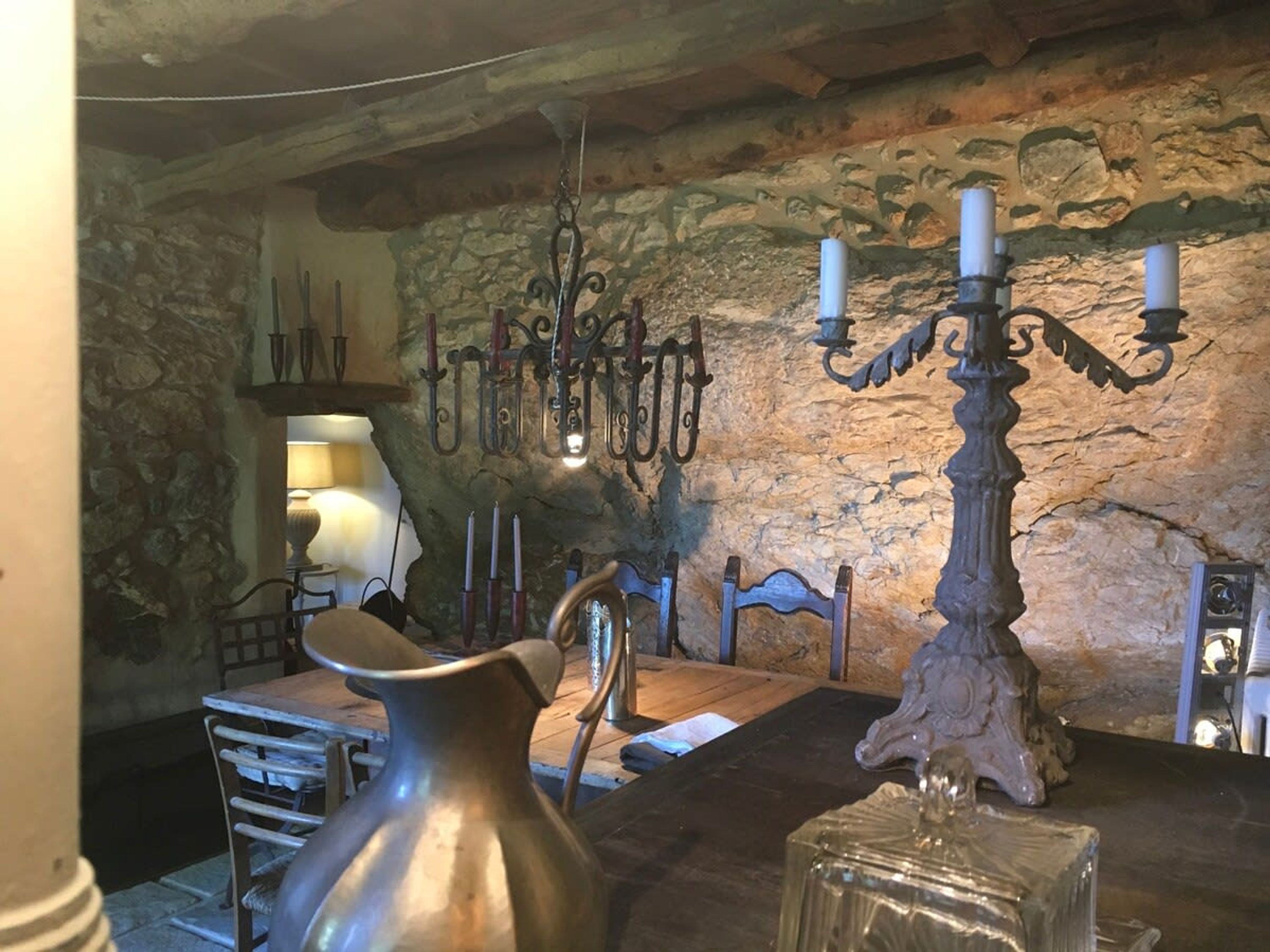 Une pièce rustique avec un mur en pierre, une table en bois, un pichet en métal, un lustre et un chandelier.