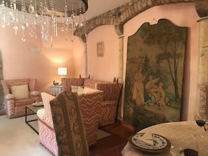Een kamer met een gestreept bankje, een eettafel met bord en wijnglas, en een groot wandtapijt.
