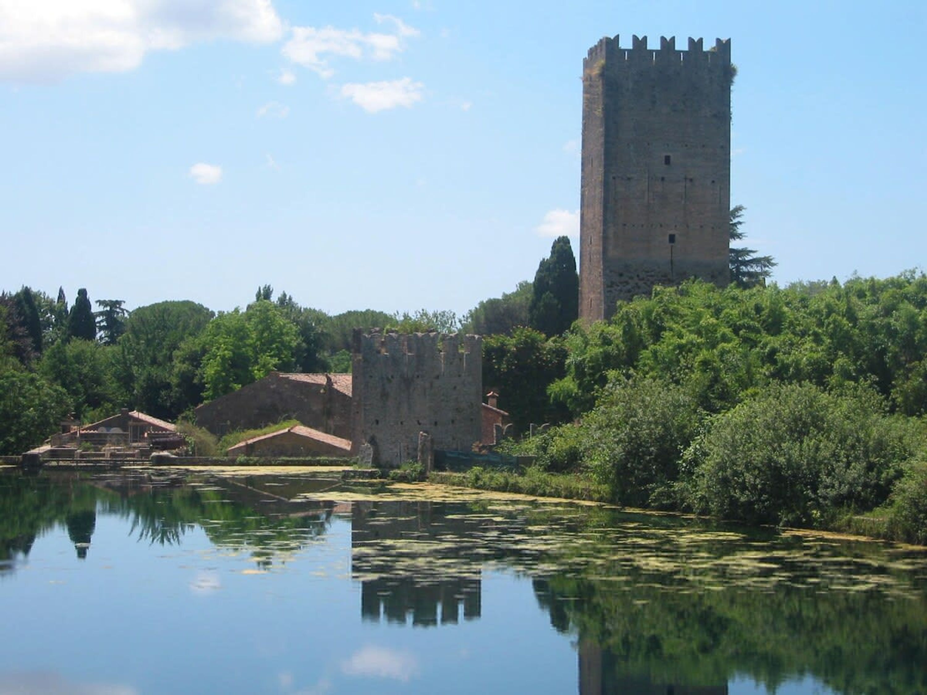 Un castillo histórico con una torre alta, rodeado de exuberante vegetación y una masa de agua tranquila que refleja la estructura.