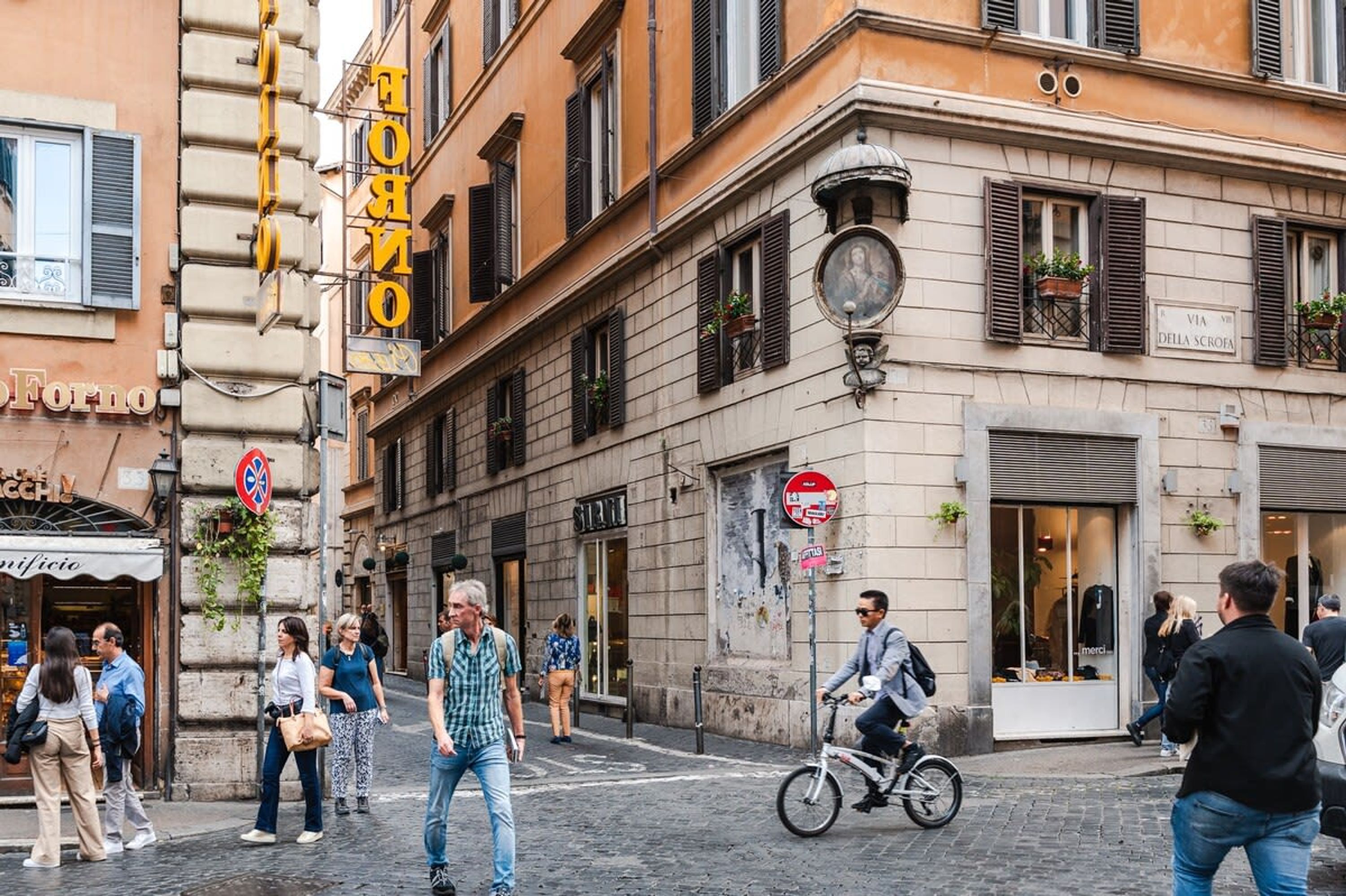 Une scène de rue avec des piétons, un cycliste et des devantures de magasins, dont une enseigne bien visible pour la « Via della Scrofa ».