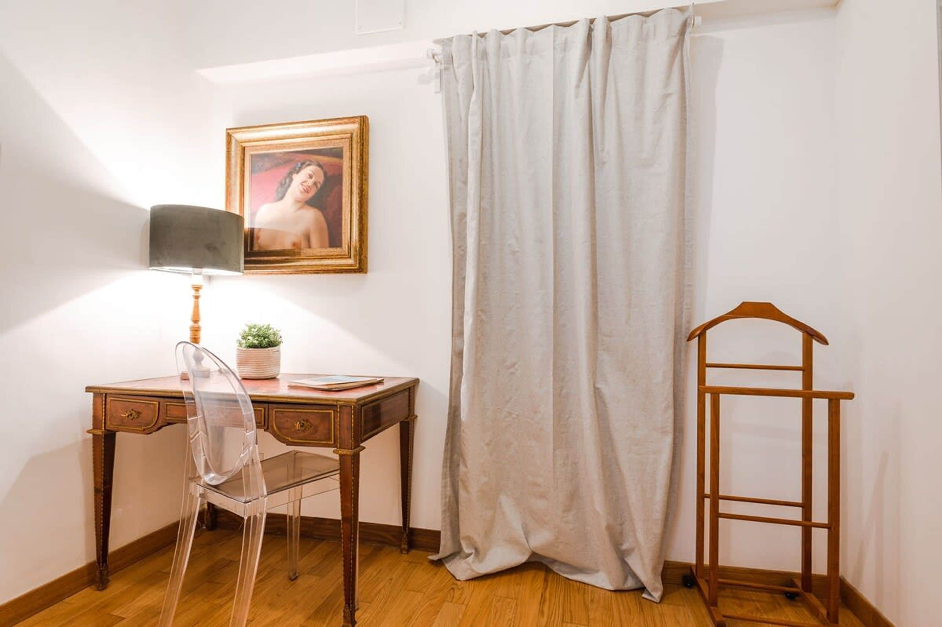 Una habitación con un escritorio de madera, una silla transparente, una lámpara, un cuadro enmarco, una cortina y un perchero de madera.