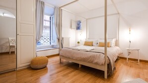 Ein Schlafzimmer mit einem Himmelbett, einem Fenster mit Vorhängen, einem Heizkörper und einem kleinen runden Pouf.