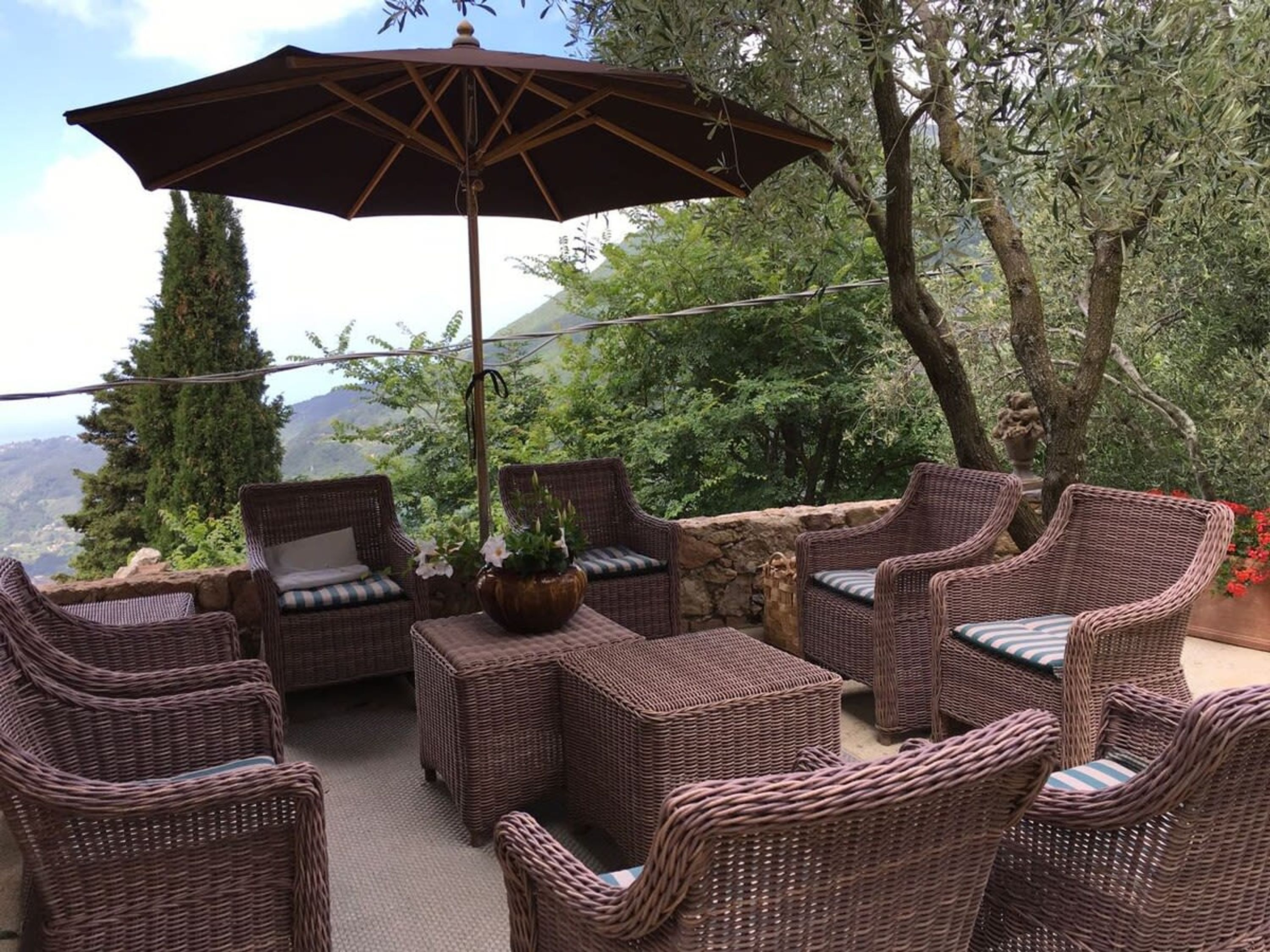 An outdoor seating area with wicker furniture and a large umbrella, surrounded by greenery and overlooking a scenic landscape.