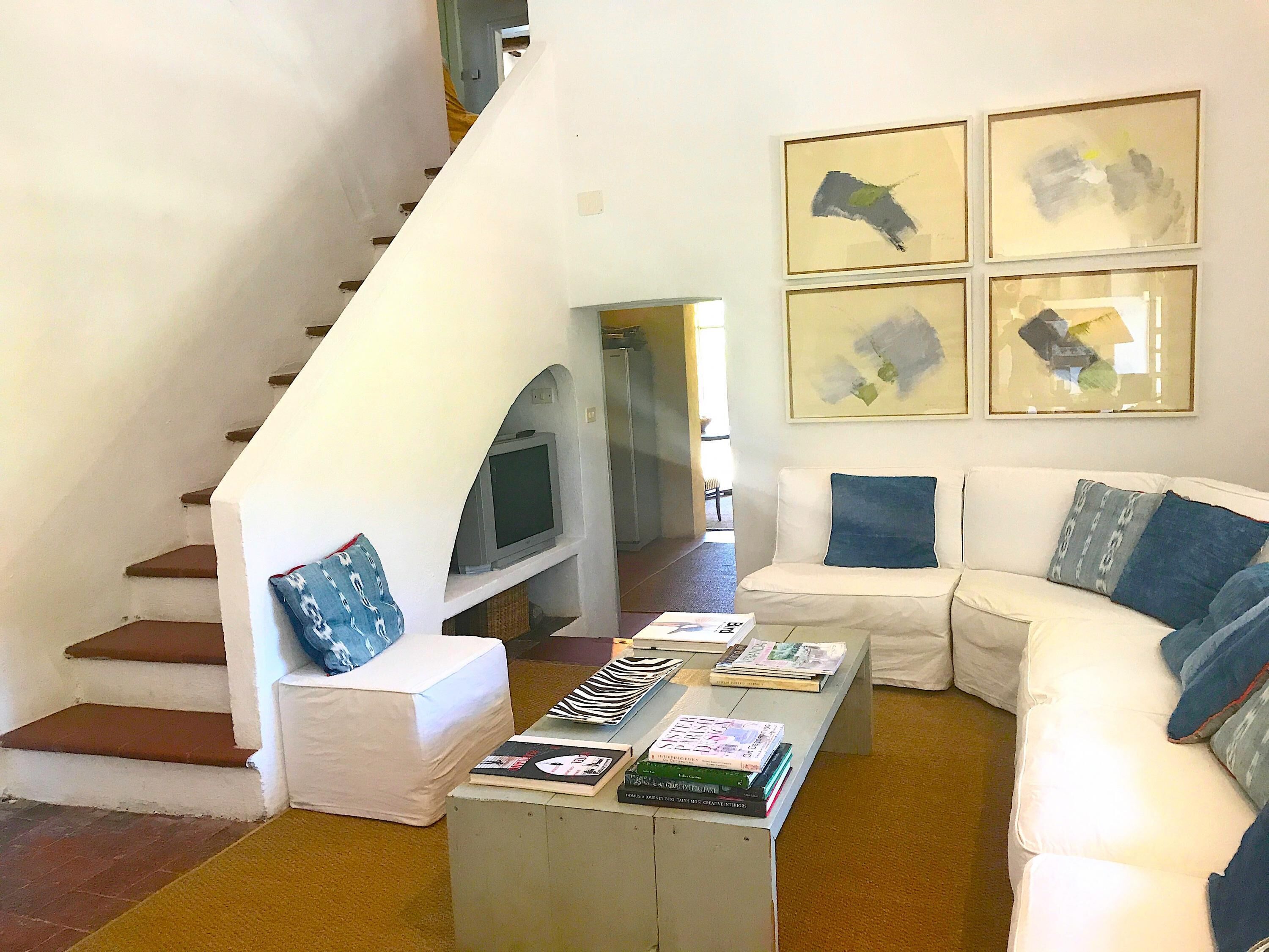 A living room with a white couch, a coffee table with books, a TV, and a staircase.