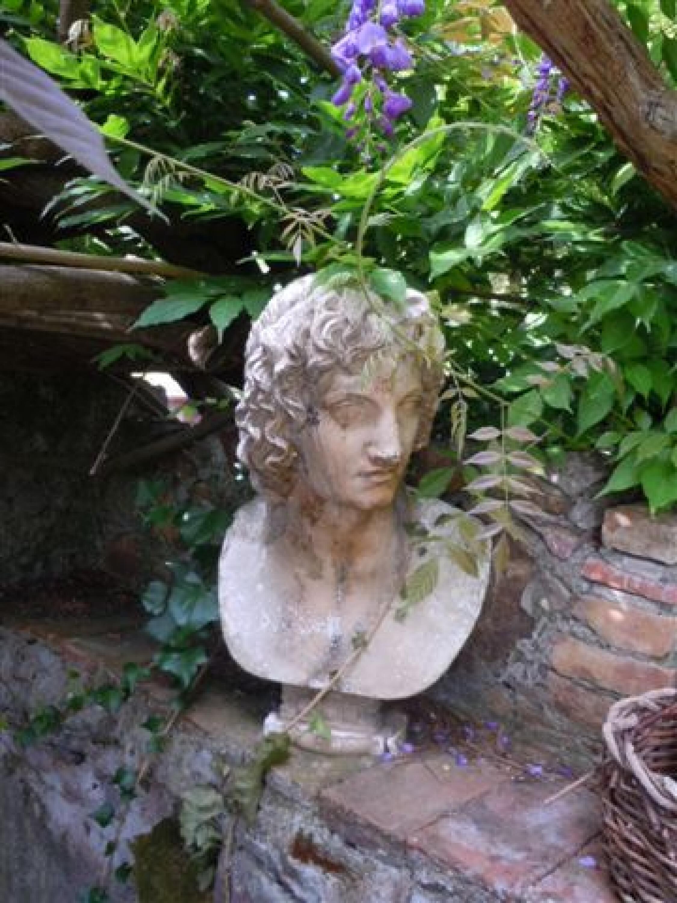 Eine Steinskulptur eines Büstenbildnisses in einem Garten, umgeben von üppigem Grün und einem Weidenkorb in der Nähe.