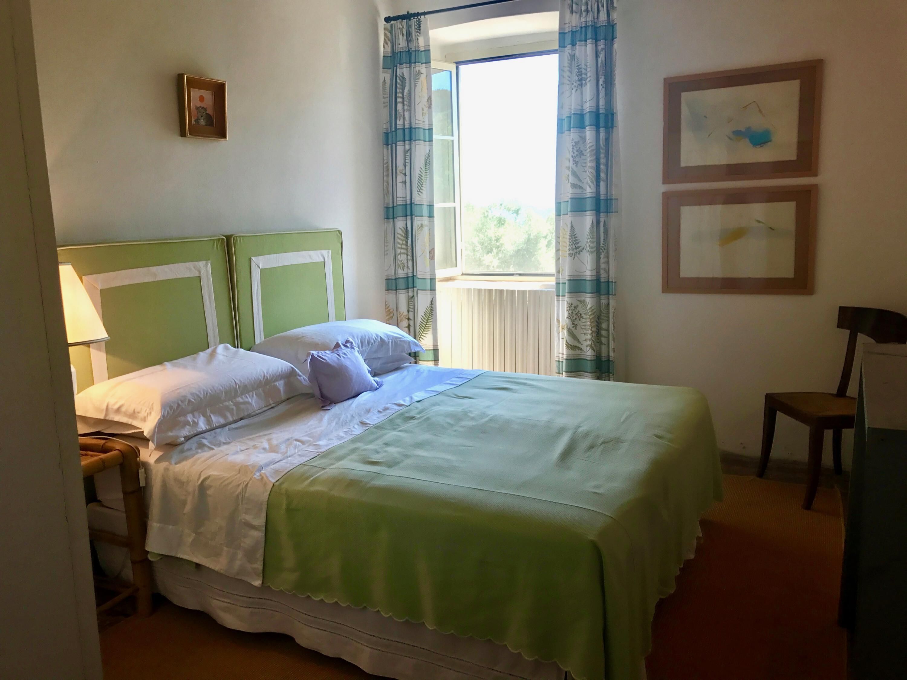 Una cama bien hecha con cabecera verde, ropa de cama blanca y una ventana con cortinas. Hay dos cuadros en la pared y una silla al lado.