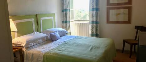 Una cama bien hecha con cabecera verde, ropa de cama blanca y una ventana con cortinas. Hay dos cuadros en la pared y una silla al lado.