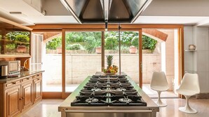 A modern kitchen with a central gas stove, wooden cabinets, and a large sliding glass door leading to an outdoor area with potted plants. - Near Rome Villa Pool Tennis Courts Perfect Family Reunion or Off-site Meeting (Grottaferrata)