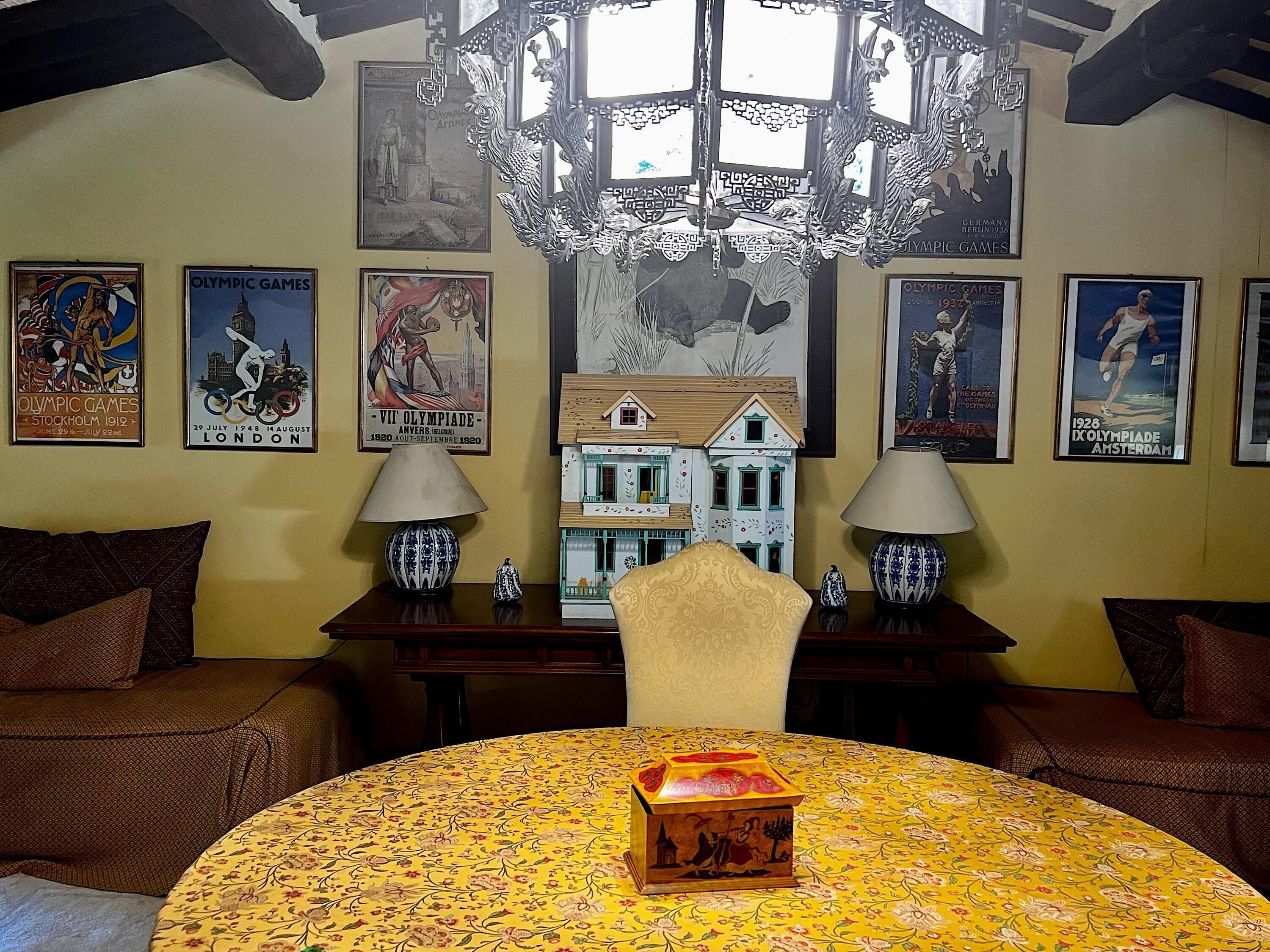 A room with a round table, two lamps, a chandelier, and Olympic Games posters on the wall.