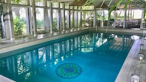 An indoor swimming pool with a geometric tile design, surrounded by large windows and shaded by umbrellas. - Villa Vinarte Elegant Home 2 Pools Tennis spa Winery Exclusively for you (Castiglione in Teverina)
