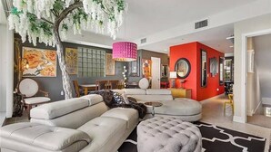 Ein modernes Wohnzimmer mit einem weißen Sofa, einem runden Hocker und einem Baum mit weißen Blüten. Eine rote Akzentwand und eine Pendelleuchte sind ebenfalls vorhanden.