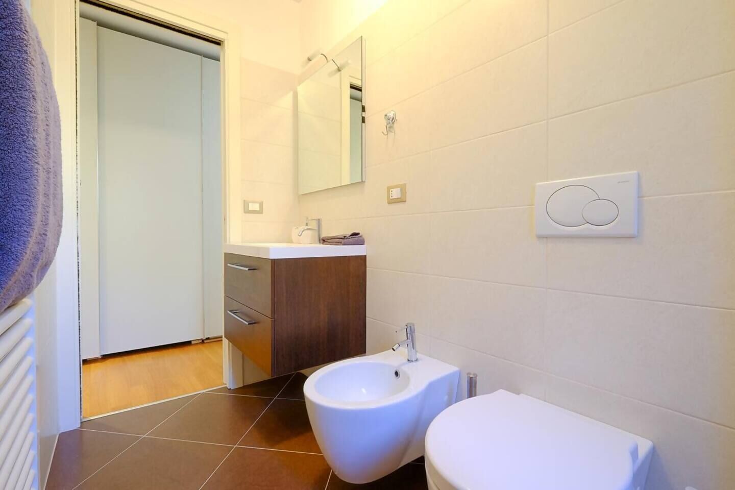 Et moderne badeværelse med en hvid vask, toilet og bidet. Et spejl er monteret over vasken, og der er en håndklædeholder på væggen.