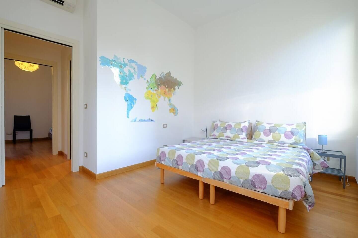 Ein Schlafzimmer mit Holzboden, einem Bett mit Punktenmuster-Decke, einer Weltkarte an der Wand und einem kleinen Tisch mit Lampe.