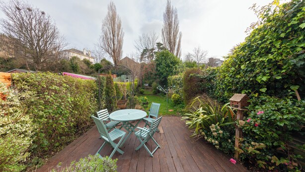 A wooden deck with a round table and chairs, surrounded by lush greenery and a birdhouse.