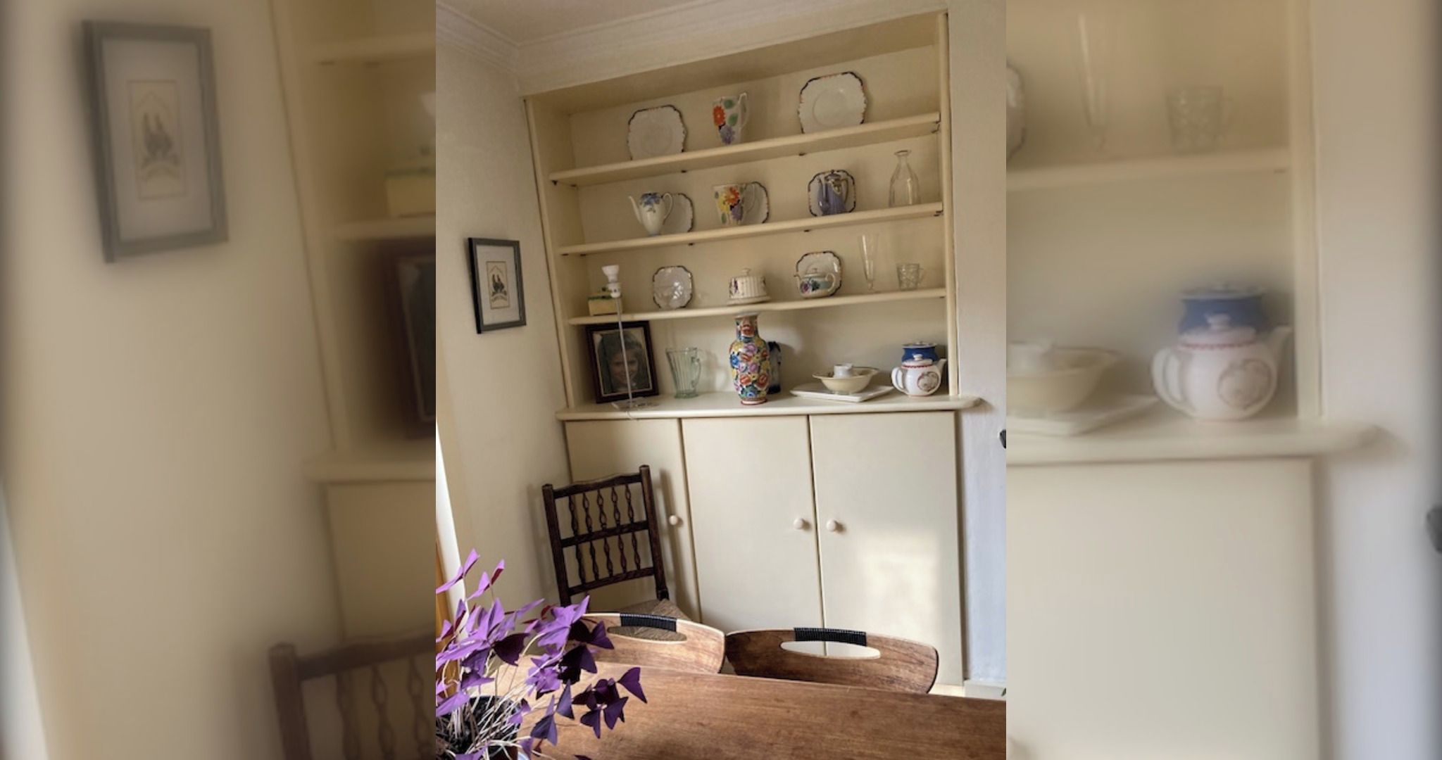 A dining area with a wooden table, a vase with purple flowers, and a cabinet with shelves of dishes.
