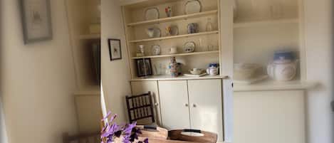 Un comedor con una mesa de madera, un jarrón con flores moradas y un aparador con estantes llenos de vajilla.