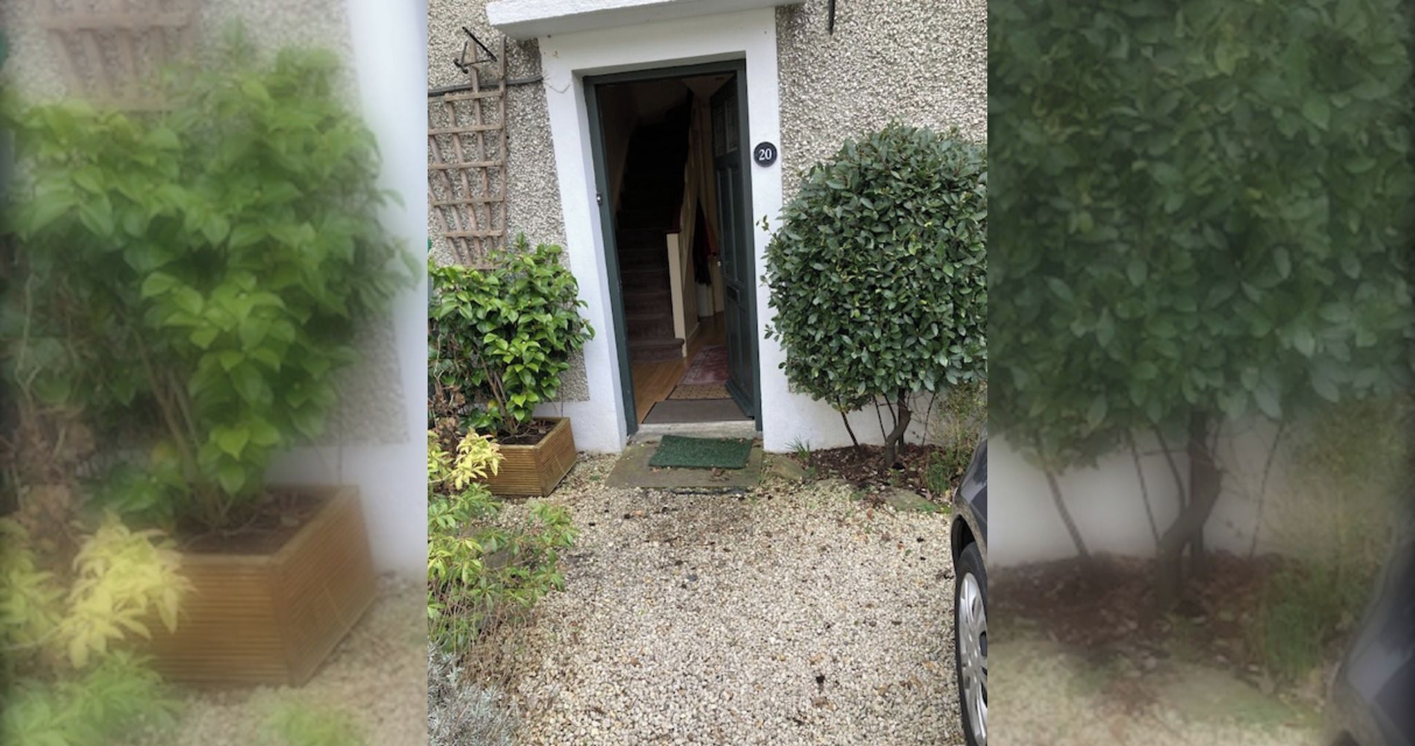 A residential entrance with a gravel driveway, a wooden planter, and a small green mat at the door.